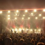Banda se apresenta em um palco iluminado diante de uma plateia durante um show de rock.
