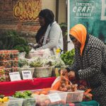 Duas mulheres organizam frutas e hortaliças em uma barraca de feira ao ar livre.