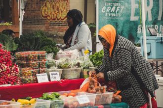 Duas mulheres organizam frutas e hortaliças em uma barraca de feira ao ar livre.