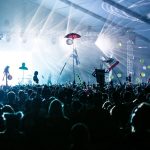 Público acompanha um show ao vivo sob luzes coloridas e elementos decorativos no palco.