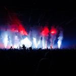 Público acompanha um show ao vivo diante de um palco iluminado por fumaça e luzes coloridas.