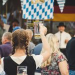 Público reunido em uma Oktoberfest ao ar livre, com canecas de cerveja levantadas em clima de celebração.